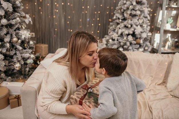 Maman est assise sur le canapé et embrasse son fils dans la pièce près du sapin de Noël