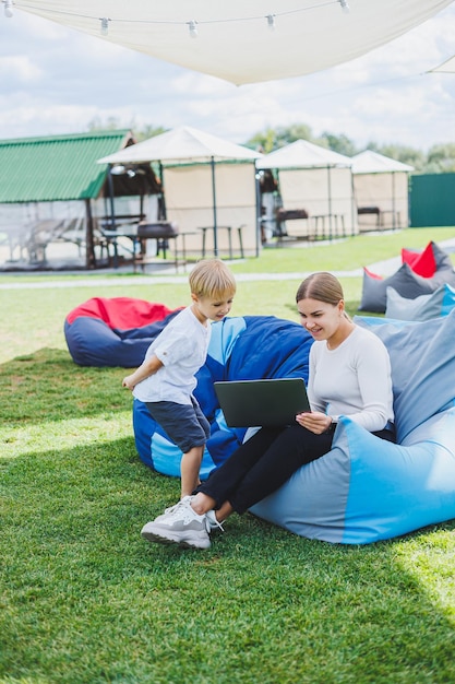 Maman est en congé de maternité Charmante jeune femme avec un petit fils travaillant à l'extérieur dans un parc d'été avec un ordinateur portable assis sur une chaise sur l'herbe verte