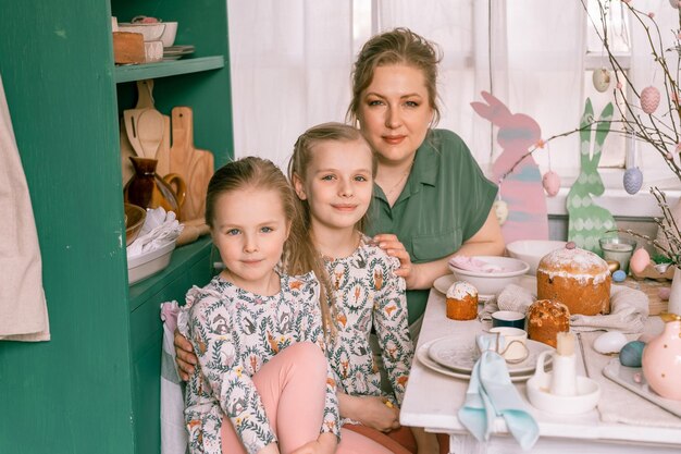 Une maman de famille heureuse avec de petites filles s'amusent au printemps pendant les vacances de Pâques à la maison dans un cadre de cuisine et décore un paysage de table avec des gâteaux de Pâques, une boulangerie et des œufs peints pour le déjeuner ou le dîner