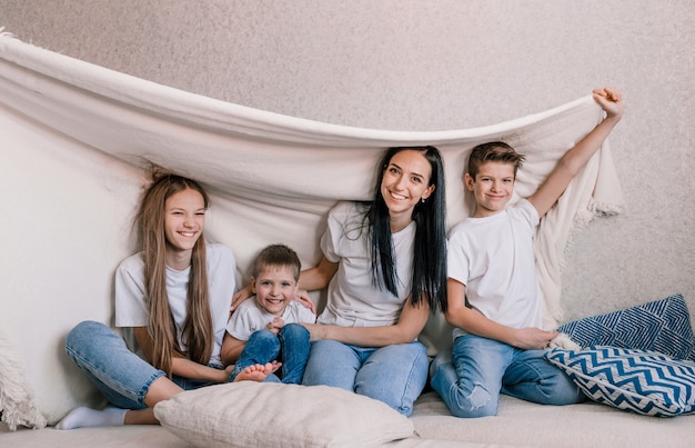 Maman de famille heureuse et petits enfants rient joyeusement assis sur le canapé recouvert d'une couverture blanche. week-end de plaisir en famille