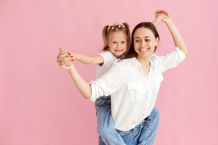 Maman femme en vêtements légers s'amuse avec une jolie petite fille. Petite fille assise sur le dos de la mère isolée sur fond rose pastel. portrait en studio.