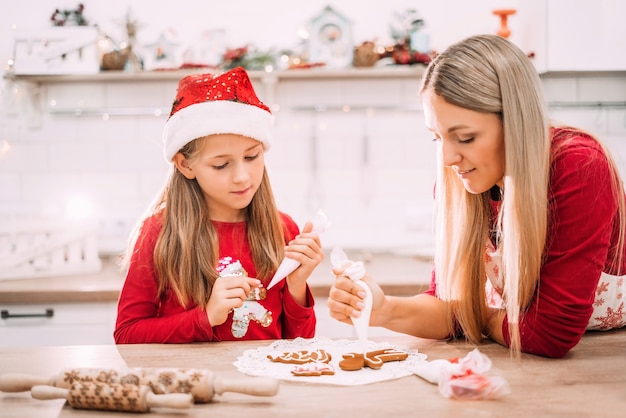 Maman et fille dans le glaçage de la cuisine sur les biscuits de pain d'épice en chandails rouges et chapeaux de Santa avec des lumières en arrière-plan