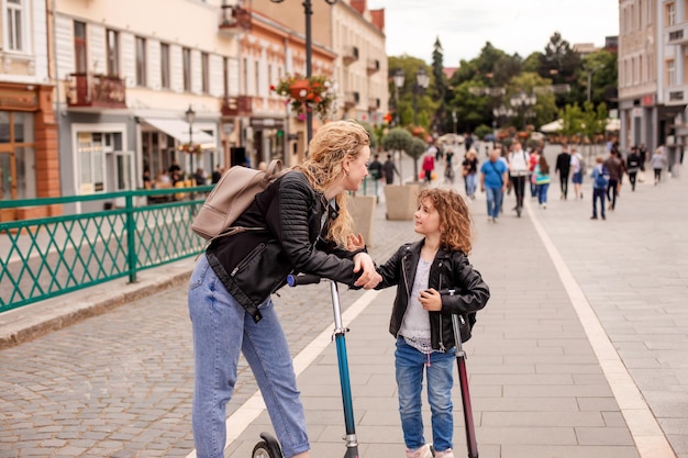 La maman et la fille heureuses se tiennent avec des scooters dans la ville