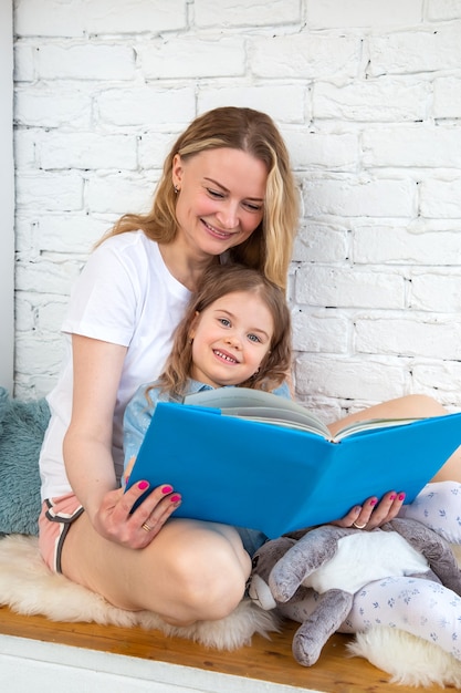 Une maman heureuse lit un conte de fées joyeux dans un livre à sa fille et sourit en étant assise dans le salon