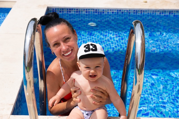 Une maman heureuse en maillot de bain rouge avec son petit fils joue à l'extérieur, sur fond de piscine aux eaux bleues.