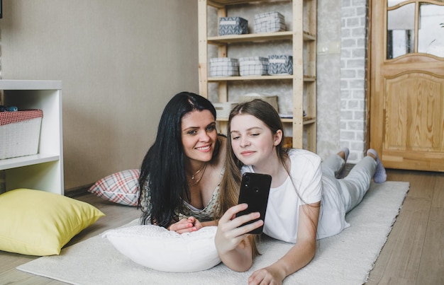 Une maman joyeuse regarde une vidéo sur son téléphone avec sa fille en train de rire allongée ensemble sur des oreillers moelleux