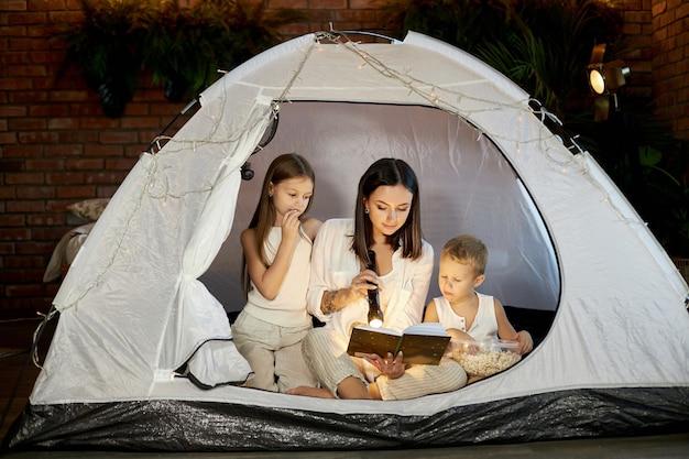 Maman lit aux enfants une histoire au coucher assis dans une tente à la maison.