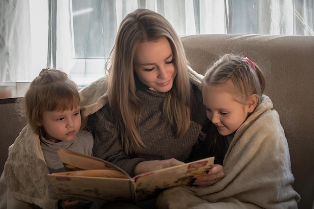 Maman à la maison sur le canapé lit un livre à ses petites filles Famille développant des loisirs à la maison