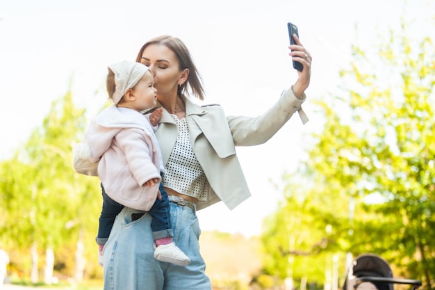 Maman mignonne avec une petite fille dans ses bras fait selfie sur smartphone dans le parc