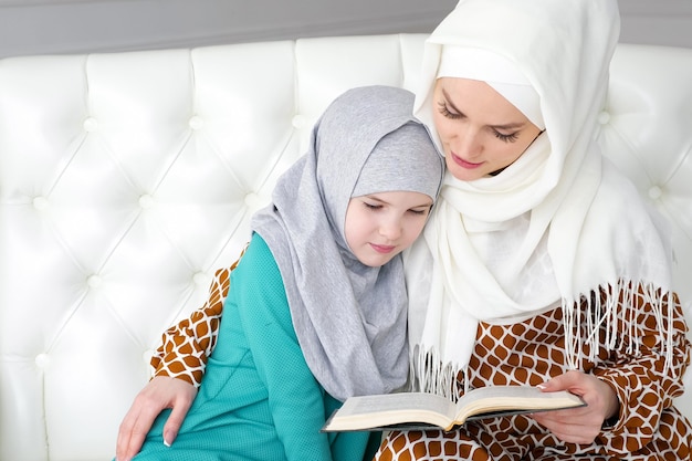 Une maman musulmane en hijab blanc et vêtements traditionnels lit pour sa petite fille un livre et la câline assise sur le canapé à la maison dans un intérieur moderne blanc.