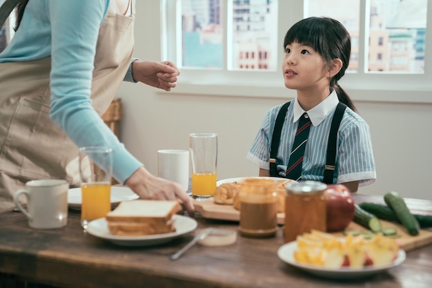 maman non reconnue en tablier debout à une table en bois avec un repas sain. jolie petite fille en uniforme regardant la mère attendant un délicieux petit déjeuner le matin avant l'école. doux moment en famille.