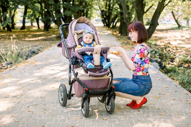 Maman nourrit à la cuillère l'enfant qui marche avec une poussette dans le parc.