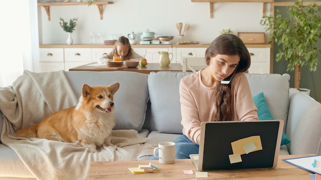 Maman occupée à essayer de travailler à distance depuis la maison. En arrière-plan, sa fille est assise à un bureau, tandis que sa mère travaille avec un ordinateur portable et a une conversation d'affaires au téléphone.