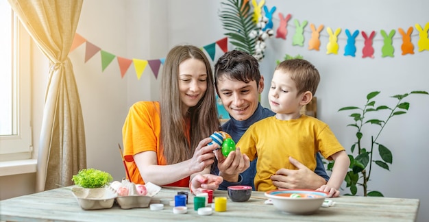 Maman papa et petit fils dans des vêtements lumineux colorent des œufs dans une pièce décorée Concept de préparation familiale pour l'ambiance printanière festive de Pâques