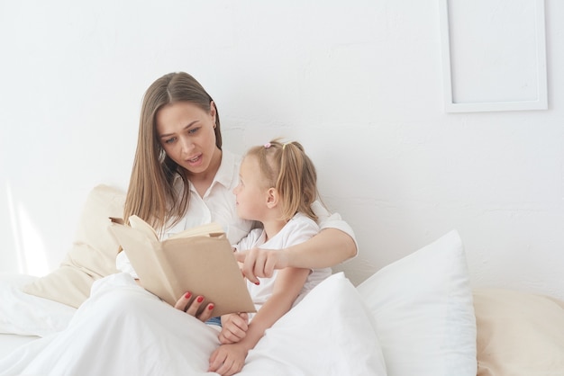 Maman et petite fille en gros plan se reposent dans un lit confortable, lisant ensemble un livre pour enfants amusant et intéressant.