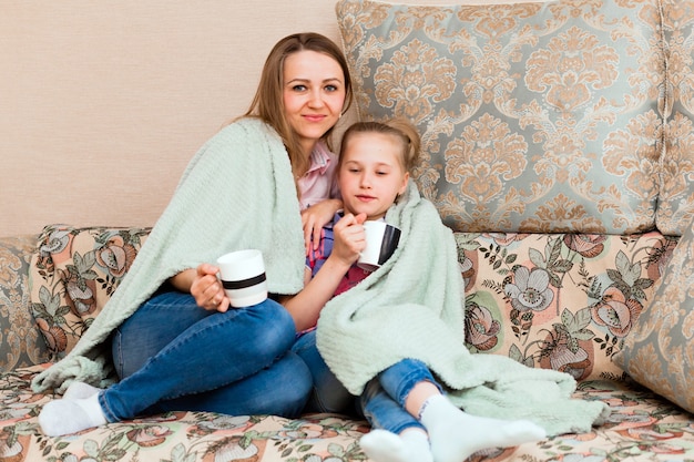 Maman et sa fille, enveloppées dans une couverture douce, s'assoient sur le canapé et boivent du thé.