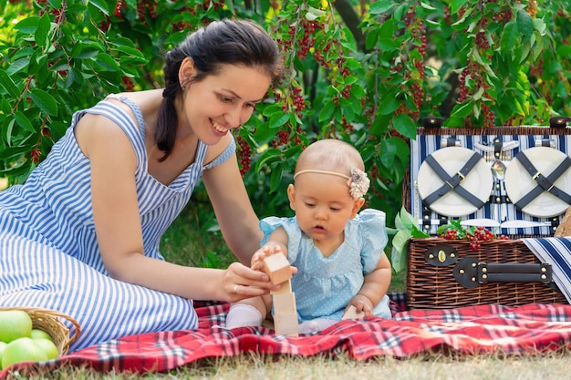 Maman et sa fille nouveau-née assemblent une tour de cubes en bois lors d'un pique-nique dans le parc. Mère et fille pendant 10 à 12 mois jouent à des jeux éducatifs sur une couverture de pique-nique