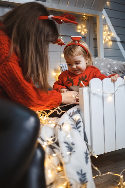 Maman avec sa fille en pulls chauds rouges saute sur le lit. Bonne maternité. Relations familiales chaleureuses. Intérieur de Noël et du nouvel an. Amour. Notion de famille.