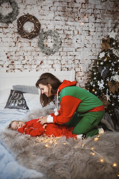Maman avec sa fille en pulls chauds rouges saute sur le lit. Bonne maternité. Relations familiales chaleureuses. Intérieur de Noël et du nouvel an. Amour. Notion de famille.