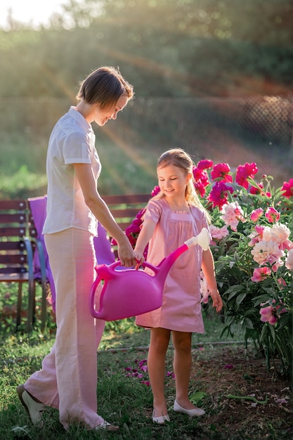 Maman et sa fille en robe rose en lin s'occupent des pivoines dans le jardin, arrosent les fleurs. Il sourit et rit parfaitement. Illuminé par les rayons du soleil couchant du soir.