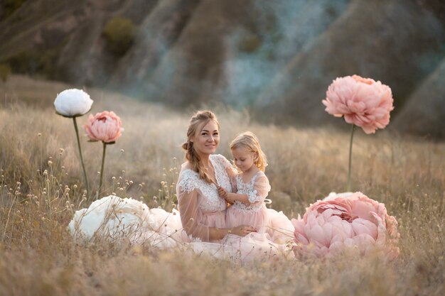 Maman et sa fille en robes de conte de fées roses sont assises dans un champ entouré de grandes fleurs décoratives roses