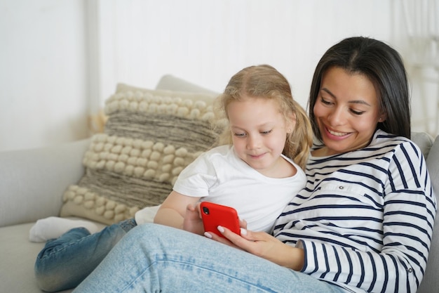 Maman et sa petite fille regardent des dessins animés sur un smartphone en profitant de leur temps libre sur un canapé ensemble