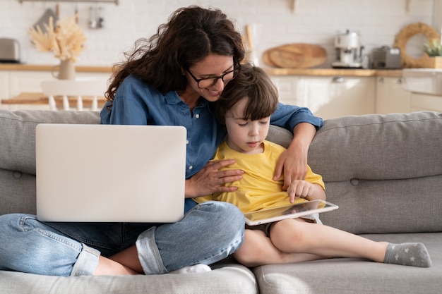 Maman et son enfant utilisent une tablette assis sur un canapé, une mère souriante aide l'enfant avec des leçons ou des jeux d'apprentissage en ligne