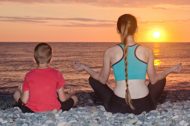 Maman et son fils méditent sur la plage en position du lotus. Vue de l'arrière, coucher de soleil