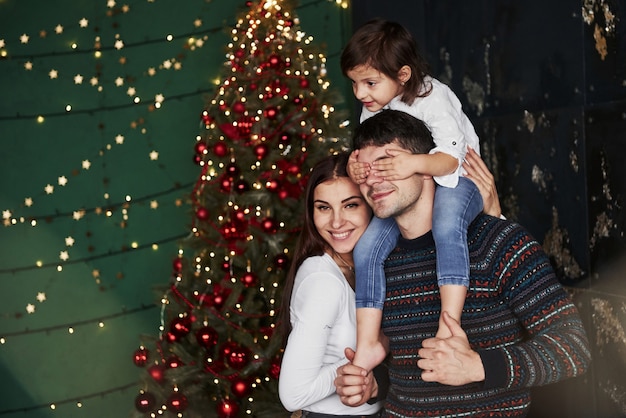 Maman sourit. La fille est assise sur les épaules du père et couvre ses yeux. Sapin de Noël décoré