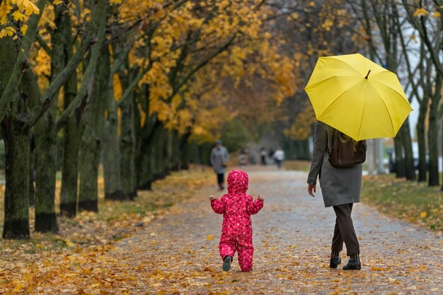 Maman sous un parapluie jaune et bébé marchent le long d'une ruelle avec des feuilles tombées à travers un parc en automne.