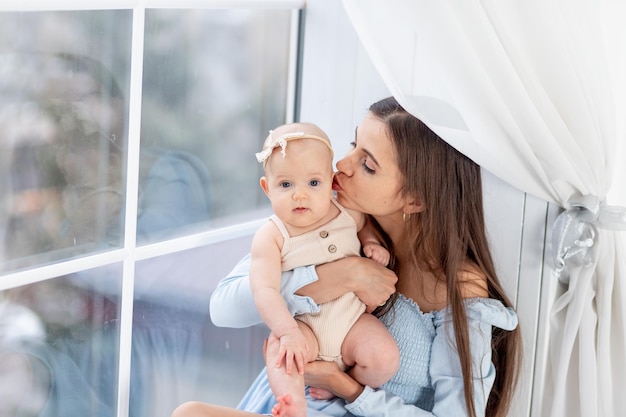 Maman tient une petite fille nouveau-née dans un costume de coton à la fenêtre de la maison l'étreignant et l'embrassant bonne maternité ou famille