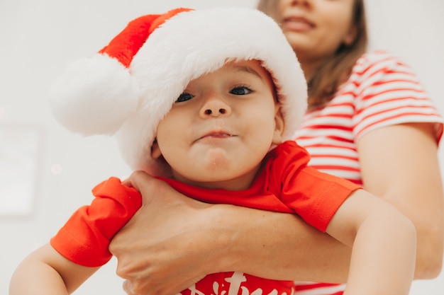 Maman tient son fils dans un costume de père Noël dans ses bras sur fond de mur avec des guirlandes. joyeux Noël
