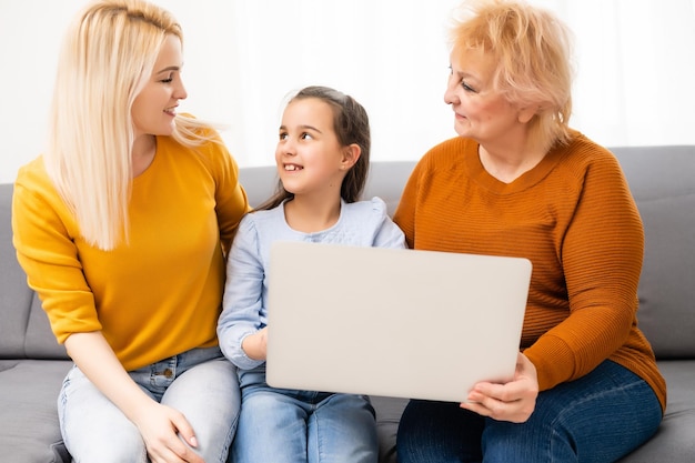 Mamie, sa fille et sa petite-fille utilisent un ordinateur portable et sourient assises sur un canapé à la maison.