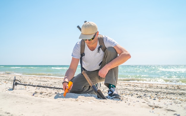 Man avec détecteur de métaux et pelle à la recherche de bijoux dans le sable à la journée ensoleillée près de la mer.