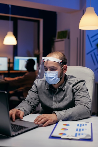 Manager et collègues faisant des heures supplémentaires avec visière contre covid-19. Homme stressé en entreprise travaillant dur pour terminer un projet portant un masque facial par mesure de sécurité en raison de la pandémie de coronavirus.