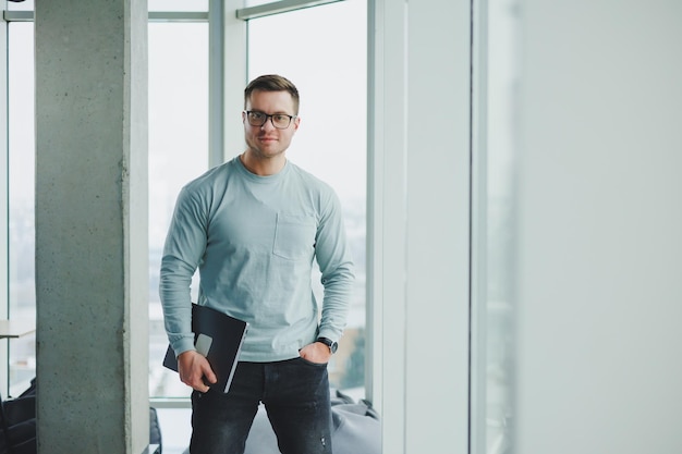 Un manager masculin moderne en vêtements décontractés se tient près de la fenêtre dans l'espace de travail avec un ordinateur portable