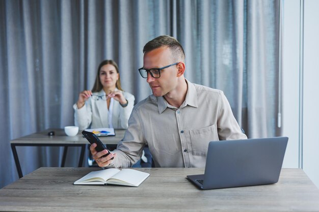 Un manager masculin parle sur un téléphone portable assis à une table avec un ordinateur et un bloc-notes Ambiance de travail dans un bureau avec de grandes fenêtres