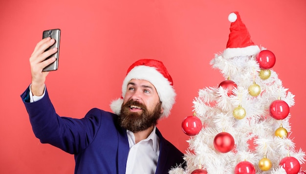 Manager santa hat utilise un smartphone. selfie près de l'arbre de Noël. Capturez un souvenir heureux. Fête du nouvel an. joyeux Noël. cadeau de recherche d'homme barbu en ligne. homme d'affaires travaillant les jours fériés. saluer ses plus proches.