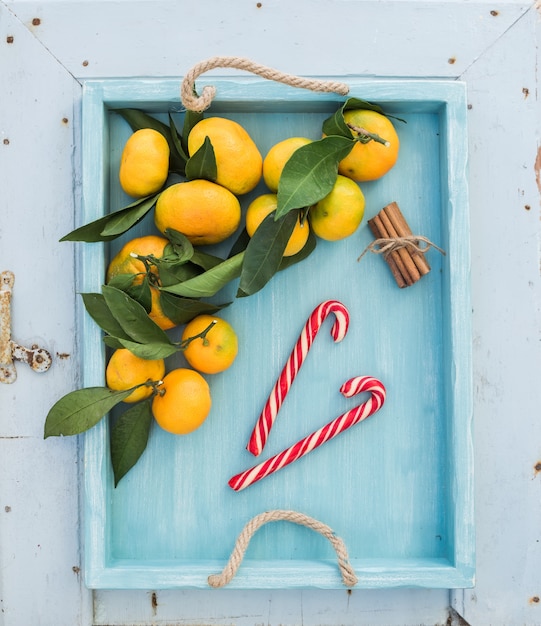 Mandarines fraîches avec des feuilles, des bâtons de cannelle et des cannes de bonbon de Noël dans un plateau turquoise sur la toile de fond en bois rustique bleue, vue de dessus.