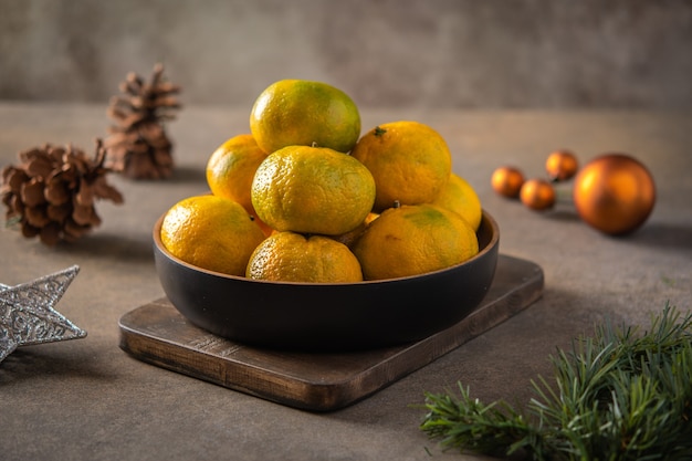 Mandarines ou mandarines et tranches mûres dans un bol en bambou sur planche de bois, à côté de la branche d'arbre de Noël, cônes et jouets