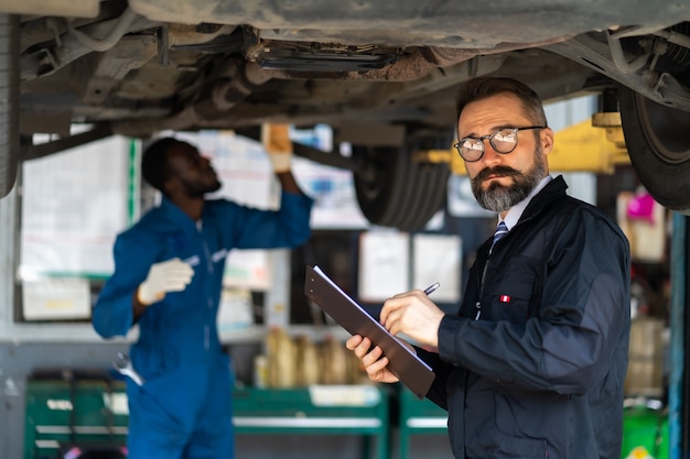 Photo une mangeoire hipster avec une barbe et un presse-papiers de liste de contrôle parlent à un mécanicien d'expertise travaillant sous un véhicule dans une station-service automobile. concept de garage d'entretien automobile et de service automobile.