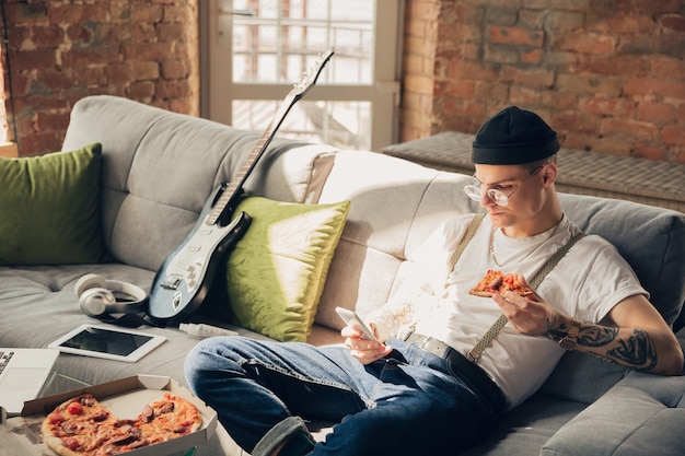 Manger des pizzas. Homme étudiant à la maison pendant les cours en ligne, école intelligente. Obtenir des cours ou une profession en étant isolé, mettre en quarantaine contre la propagation du coronavirus. Utilisation d'un ordinateur portable, d'un smartphone, d'un casque.