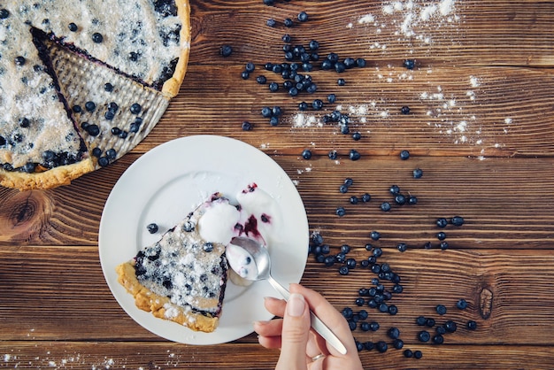 Manger une tarte aux myrtilles sur une table rustique sombre servie avec de la crème avec une cuillère. Tas de baies près du dessert. Vue de dessus.