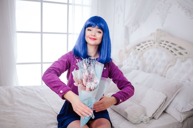 Maniaque De La Mode. Glamour Fille Synthétique Souriante, Fausse Poupée Aux Cheveux Bleus Tient Un Bouquet De Fleurs Assis Sur Le Lit Blanc.