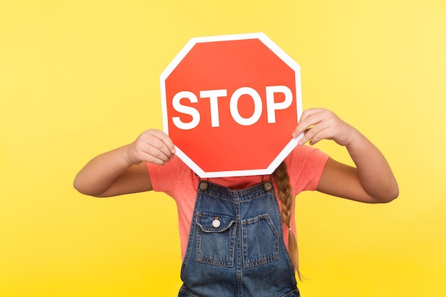 Manière interdite Petite fille en salopette en denim couvrant le visage avec le symbole d'arrêt octogonal tenant un panneau de signalisation rouge attention à la route sûre traversant la prise de vue en studio intérieure isolée sur fond jaune