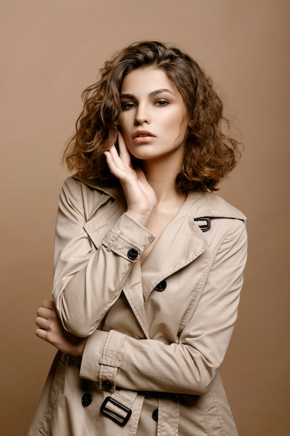 Mannequin de beauté avec une peau propre et des cheveux bouclés en manteau beige extensible avec une écharpe sur un mur beige