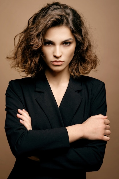 Mannequin de beauté avec une peau propre et des cheveux bouclés en veste noire sur mur beige, femme d'affaires sérieuse