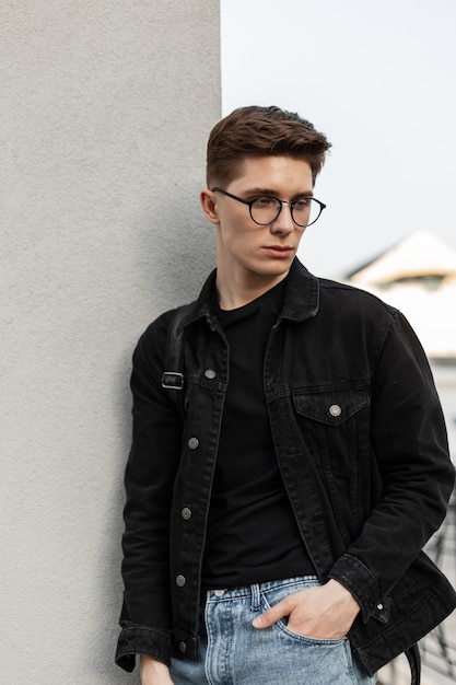 Mannequin jeune homme sérieux en jeans élégants vêtements décontractés pour jeunes dans des lunettes à la mode avec sac à dos reste près du mur en ville. Mec branché à l'extérieur. Nouvelle collection de vêtements pour hommes. Style de printemps.