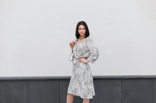 Mannequin jolie femme avec coiffure en robe de fleurs de printemps avec sac marche dans la rue près d'un mur blanc