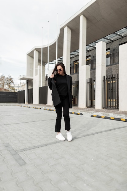 Mannequin séduisante jeune femme dans des lunettes de soleil élégantes dans des vêtements noirs à la mode de la nouvelle collection à la mode d'été se promène près d'un bâtiment moderne en ville. Belle fille en veste vintage à l'extérieur.
